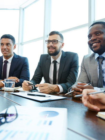 Three businessmen in suits sitting at a conference table with documents and pens.